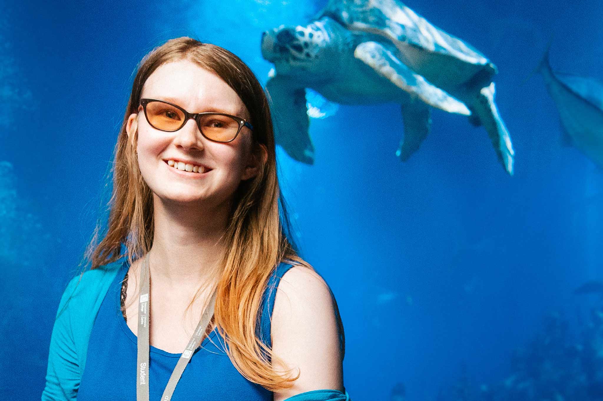Student Kiera stands smiling in a sea life and aquarium centre, with a swimming turtle behind her