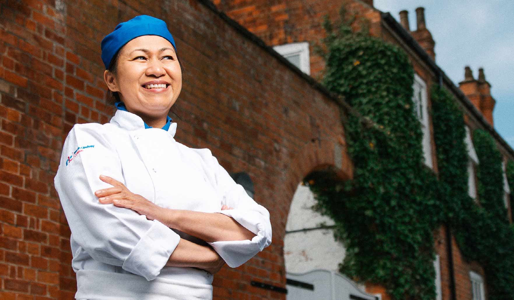 Catering student Tenie stands with arms folded and smiling widely, wearing chef uniform. Tenie is stood in front of a large brick building which appears to be a high class restaurant.