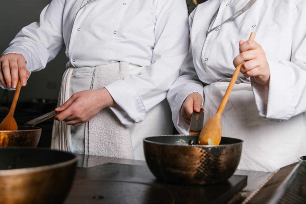 A close up image of two people in chef clothing at work in a kitchen setting. Both are using wooden spoons to mix ingredients in metal bowls set on the worktop in front of them