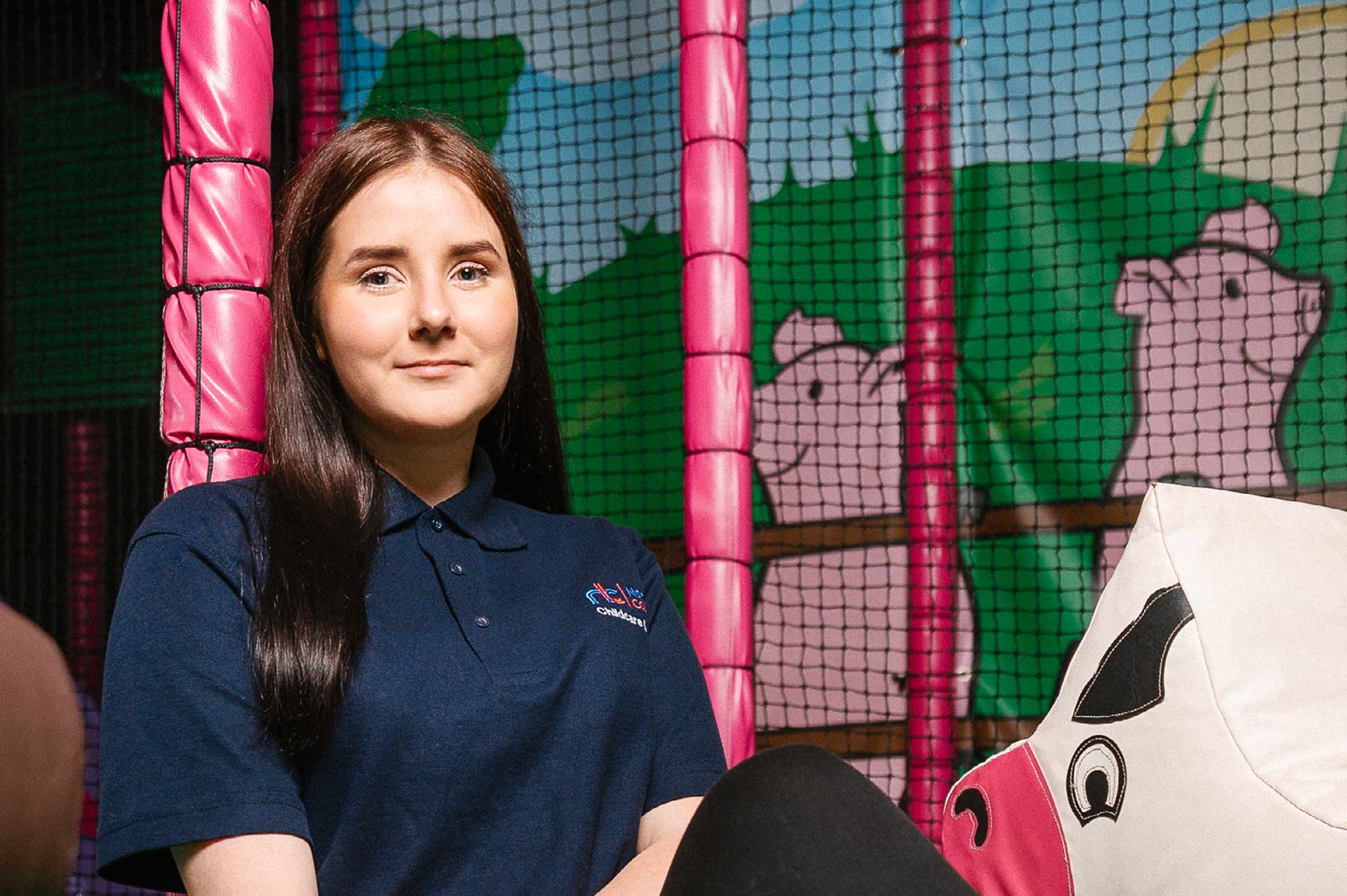 A student sits against brightly coloured soft play equipment, wearing a uniformed t-shirt and smiling to us