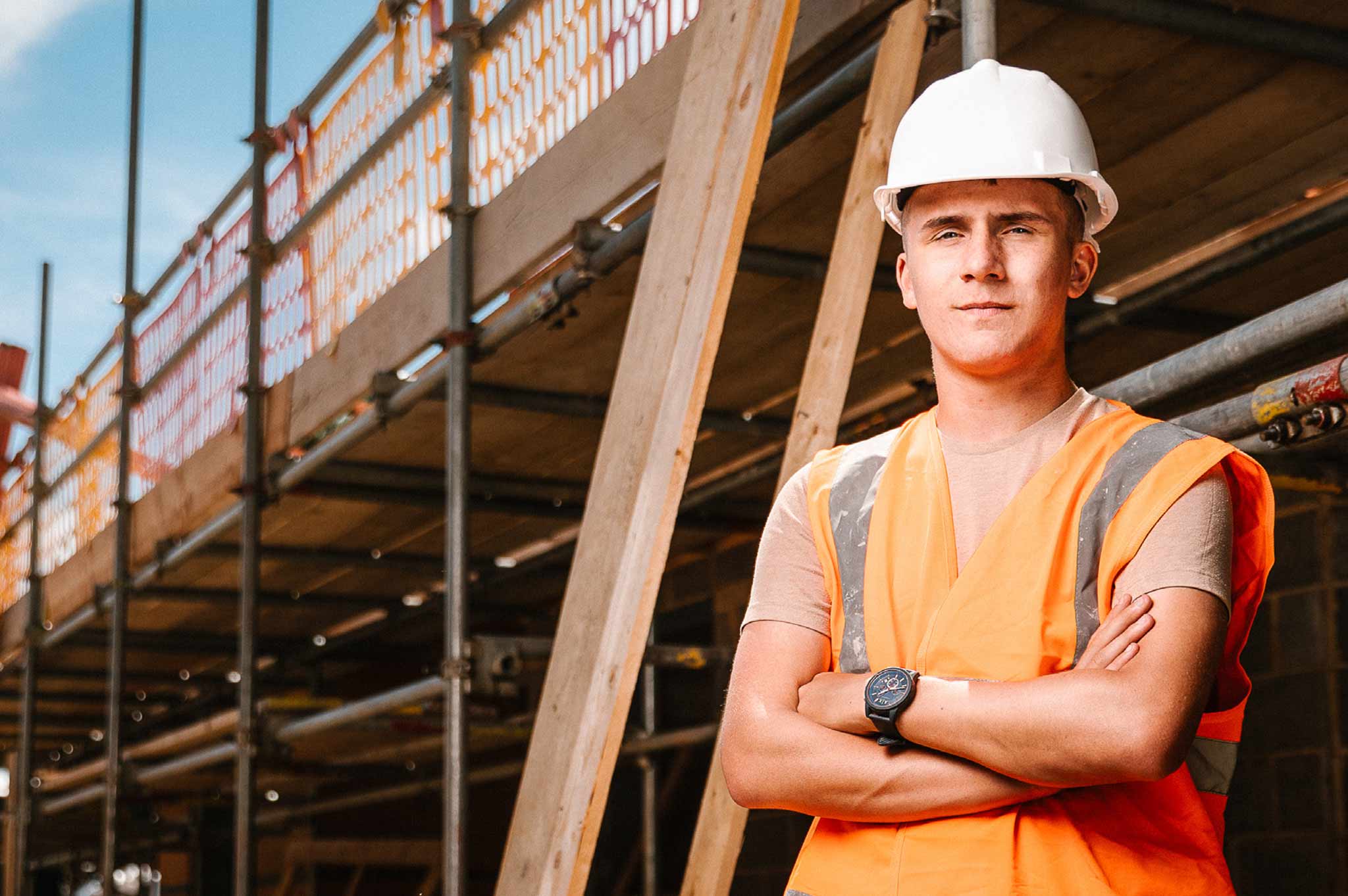 Wearing a hard hat and high visibility clothing, a student stands with folded arms against a backdrop of construction work behind.