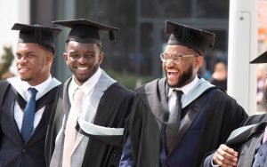 Stood outside in front of a glass fronted building, a group of smartly dressed students laugh with wide smiles after graduating. Each of them is wearing a graduate robe, hood and mortar board cap over their suits