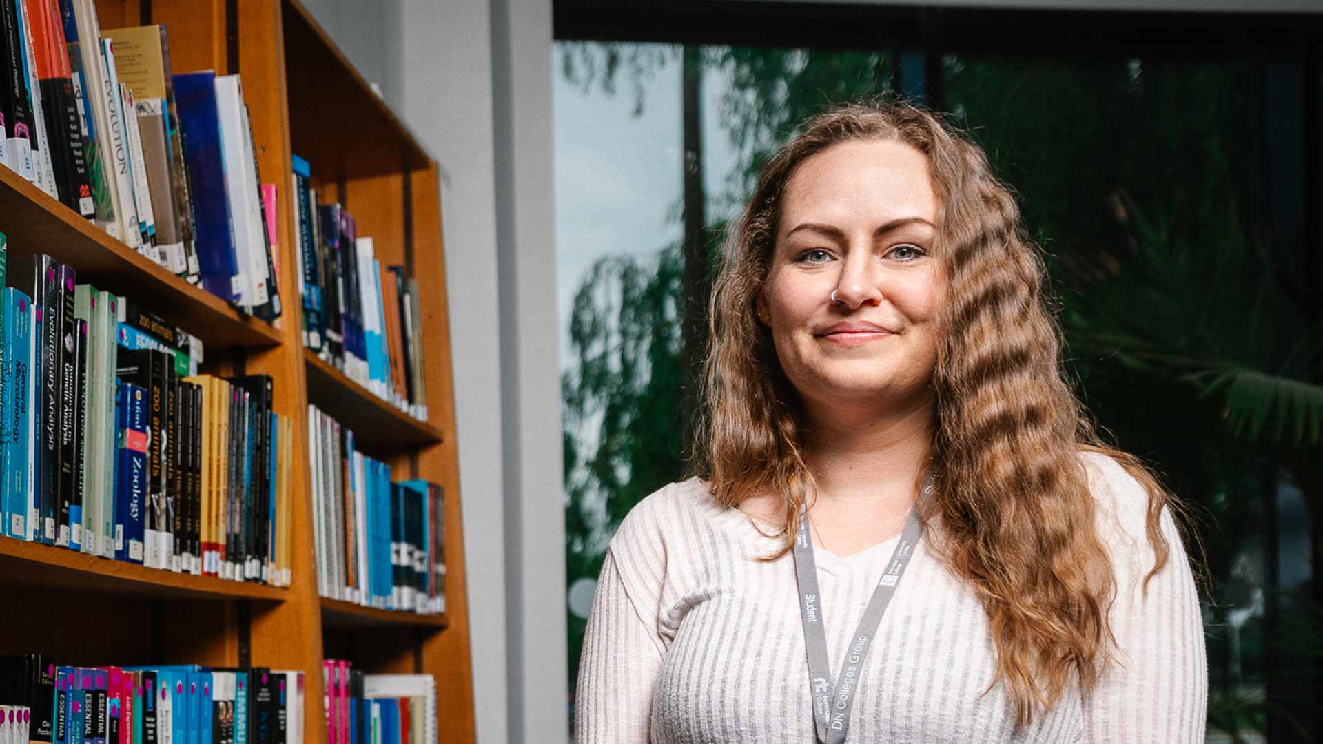 Student Karina stands smiling in front of a row of bookshelves packed with books