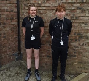 Two students wearing sports uniforms stand side by side during an Active Fusion work experience session. They stand just in front of a bare brick wall
