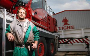 Apprentice Rici is wearing grey and emerald green overalls, leaning against a ladder with his elbow resting on it. The ladder is part of some large red machinery, the wheels of which are almost at Rici's shoulder height. They are stood in front of a building with signage which reads the company name, Tighe.