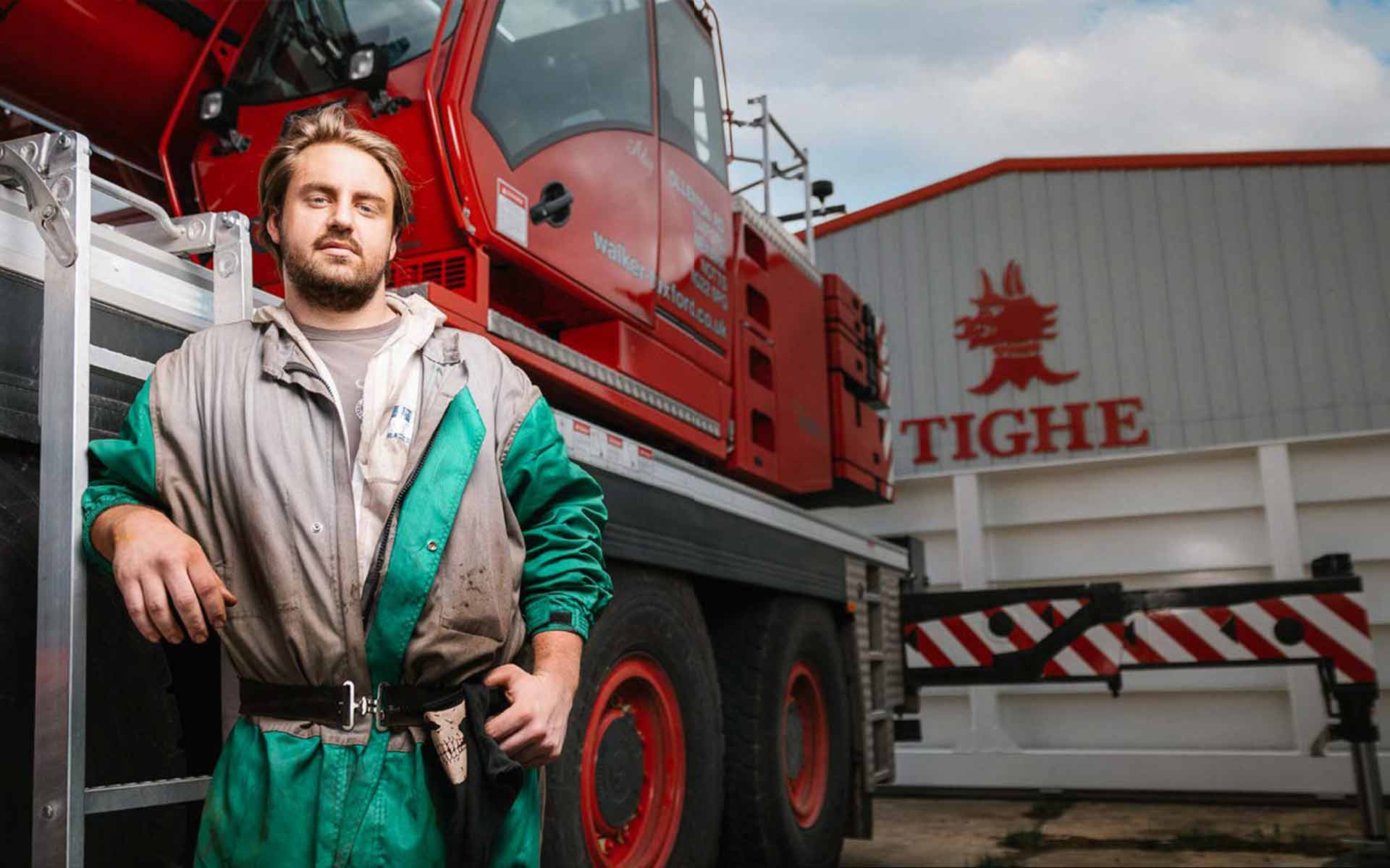 Apprentice Rici is wearing grey and emerald green overalls, leaning against a ladder with his elbow resting on it. The ladder is part of some large red machinery, the wheels of which are almost at Rici's shoulder height. They are stood in front of a building with signage which reads the company name, Tighe.