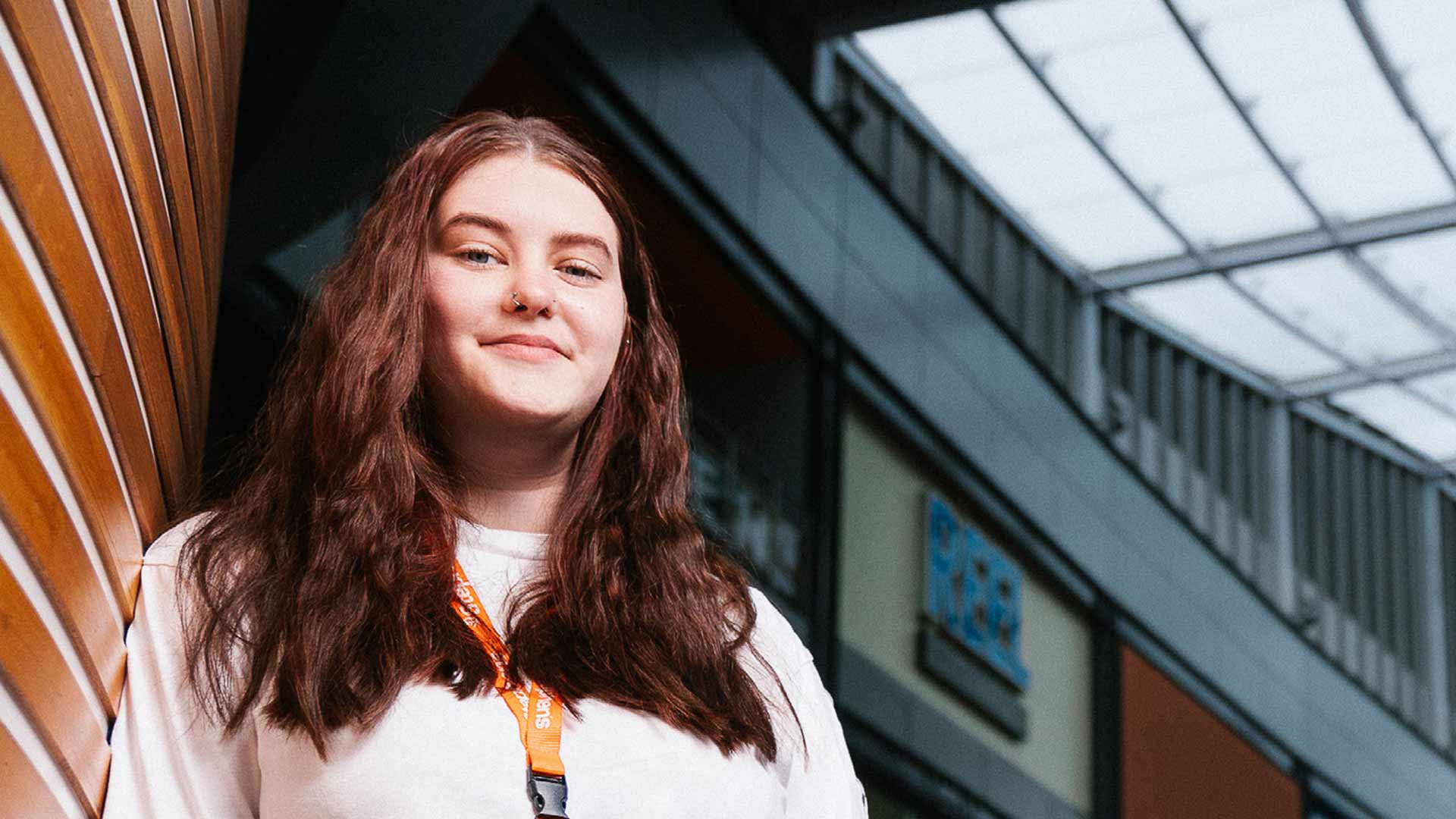 Student Rhyanna stands proud and smiling, leaning against a wooden wall in a shopping centre. A number of shop fronts are visible in the background.