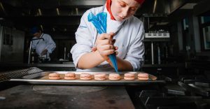 A student wearing chef uniform and a red hat holds a blue piping bag, piping cream onto a row of macarons in front of them. Surrounding them is a dark metal kitchen setting