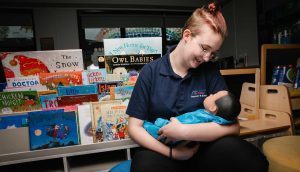 Stood in front of a range of children's books on book shelving, a student smiles at an interactive model baby doll, wrapped in a blue blanket.