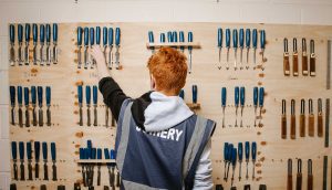A student facing away from us, wearing a blue work overall jacket, reaches to a large wooden wall containing a vast array of chisels and other woodworking tools