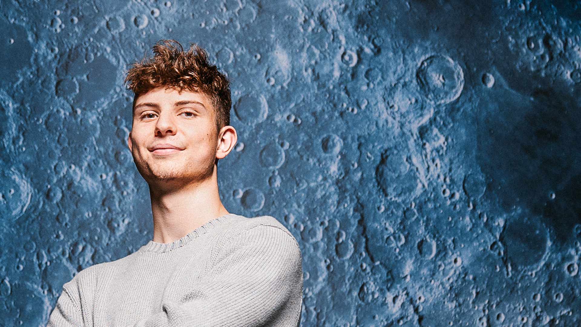 Joseph stands with arms folded in a grey sweatshirt, smiling. Behind him is a large image of a close up of the moon's surface in a blue tint
