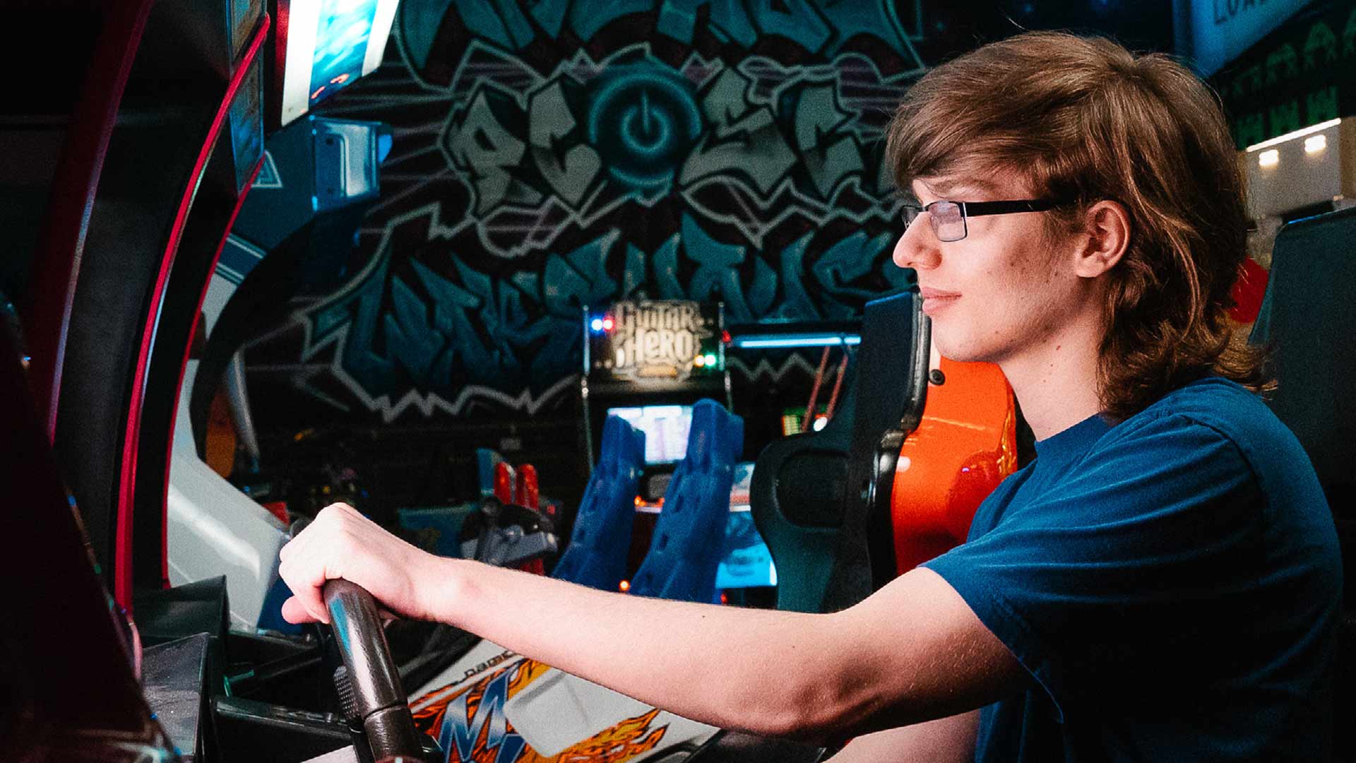 In a games arcade surrounded by multiple gaming machines, student Kenny sits at a racing car game with hands on the steering wheel. In the distance a wall at the back of the room features a stylised power button and graffiti text artwork