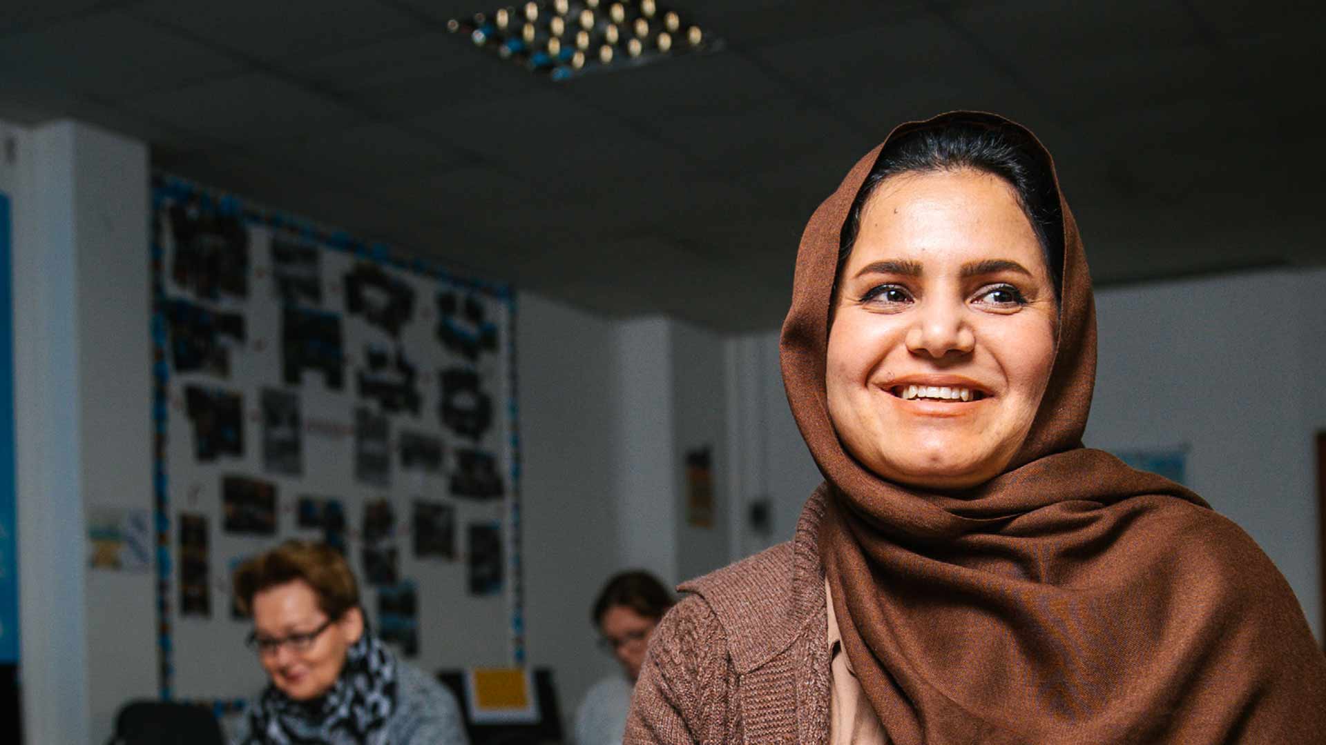 Student Rozhan sits smiling and looking off to the side. Behind Rozhan, other students are sat studying at their desks, and blurred in the background some student work is displayed on the rear wall