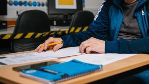 A close up image of a student wearing a blue tracksuit jacket with hands rested over various sheets of paper on the desk. A yellow pen rests casually in their hand and a notebook and phone sit on the table closer to us