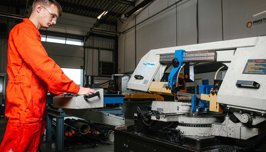 Wearing vibrant orange safety overalls, a student looks intently at a large bandsaw machine which has its blade set over a piece of metal tubing. The student is controlling the machine using a control panel set at waist height in front of them. Underneath the machine, various pieces of metal tubing can be seen in storage