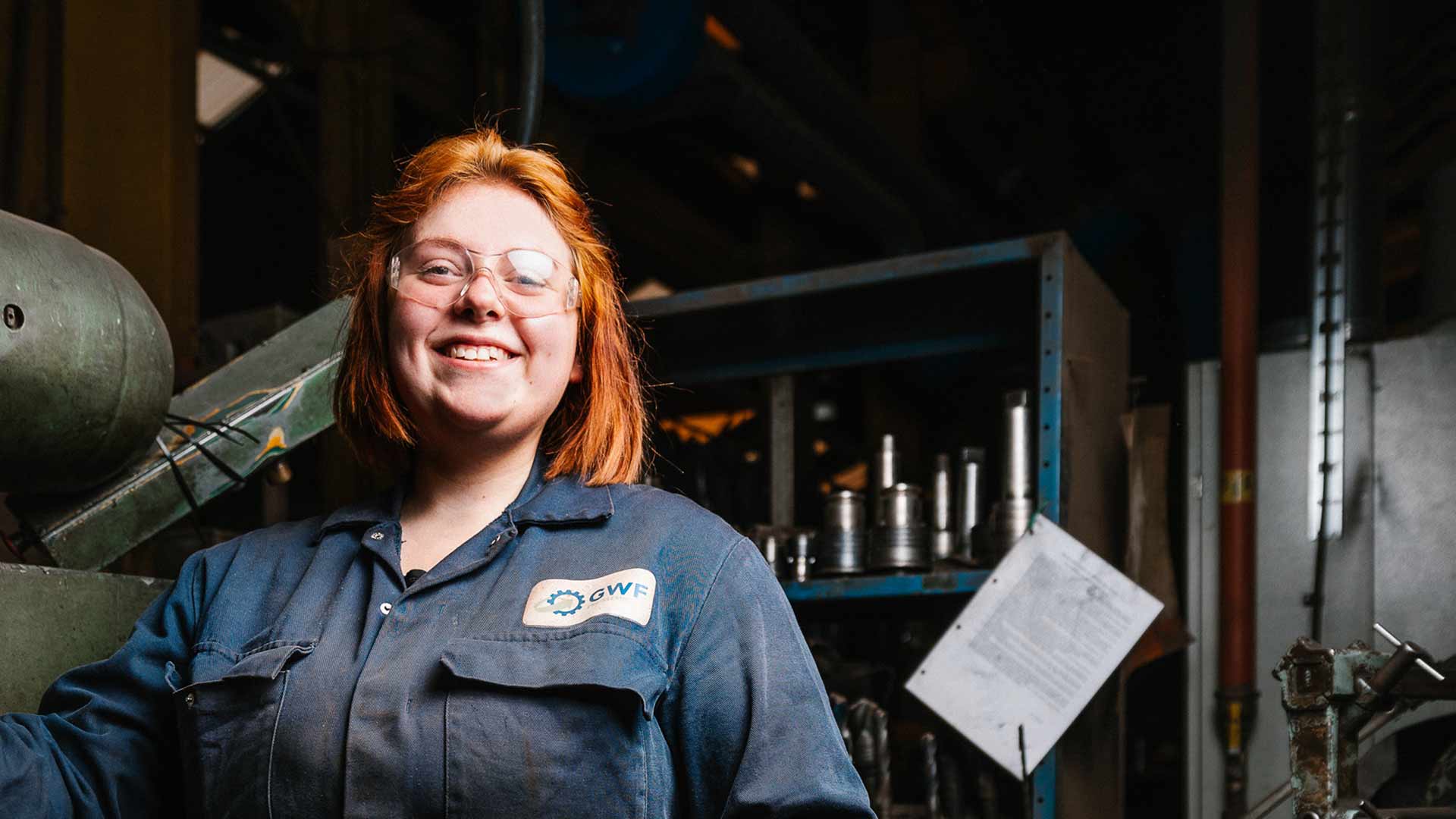 In an engineering working area, student Alisha smiles towards us, wearing safety glasses, gloves and overalls. Behind her a document on white paper is pinned to shelving which appears to contain spare equipment and tools