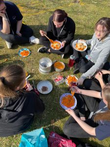 During a camping trip on a sunny day, a group of 6 students sit on the grassy ground. Each holds a plate or bowl filled with pasta and meatballs with a red tomato sauce. The empty cooking pot and stove rest in the middle of the group after being used to cook their meals