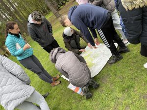 In a patch of grass surrounded by trees in the background, a group of students huddles around a large map which is placed on the floor as they plot their next moves on a Duke of Edinburgh expedition
