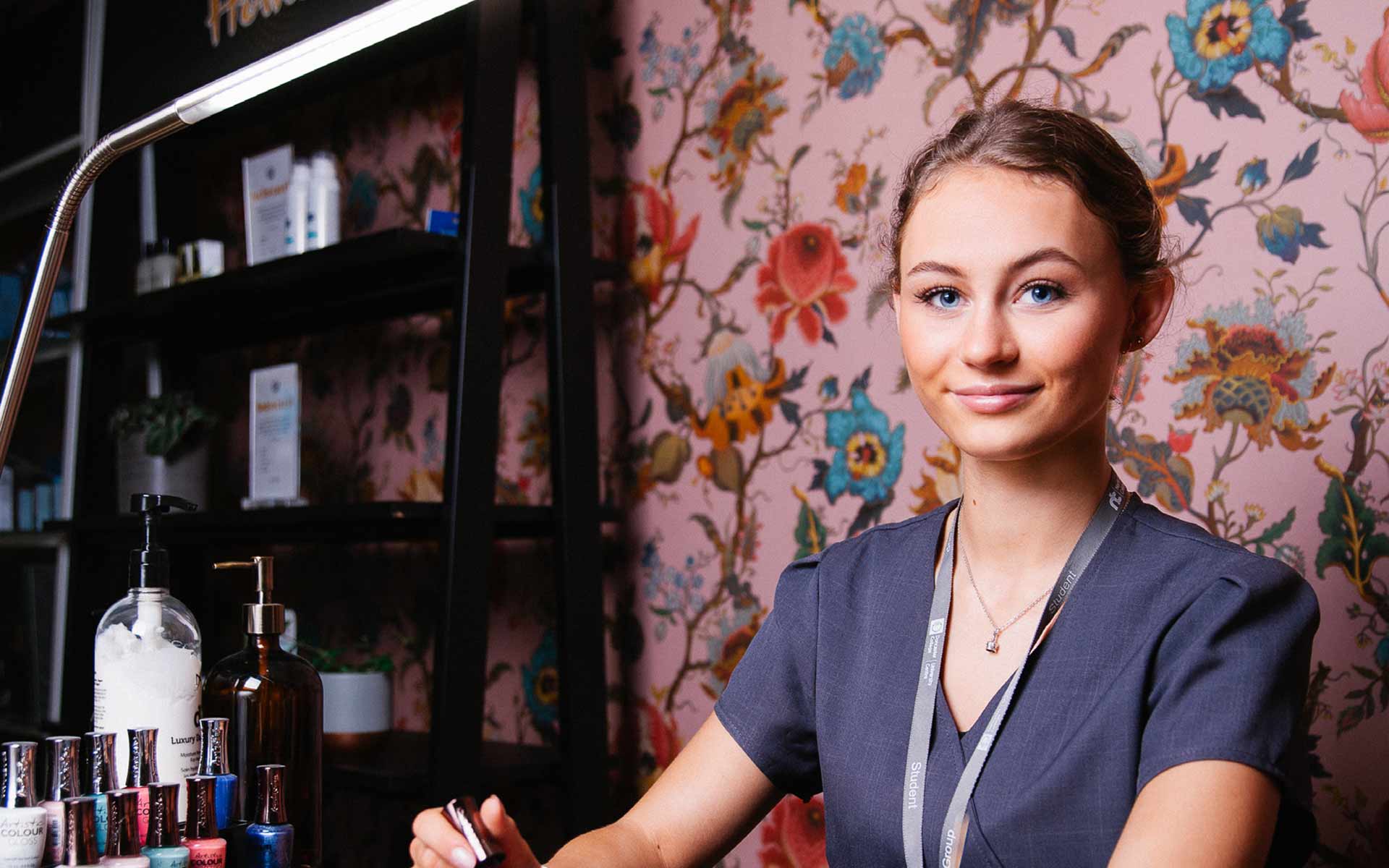 Student Freya sits in front of a pastel pink wallpaper featuring large, vibrant illustrations of flowers and branches. To her side and further into the background are a variety of make up products and items including lotions and nail polish coloured bottles. Freya wears a professional uniform and is holding the lid of a nail polish bottle in her hand