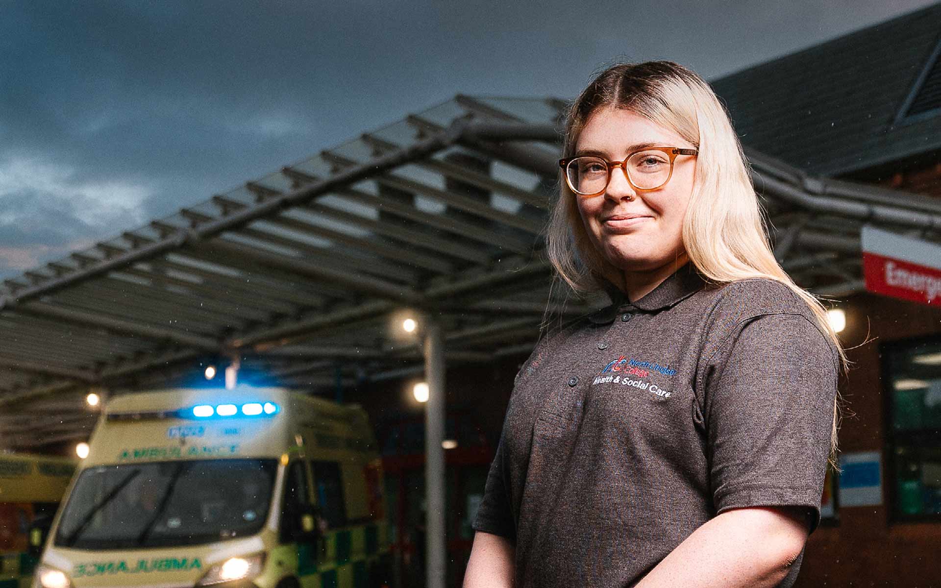 Stood outdoors at the entrance to a hospital emergency department, student Taylor smiles towards us. Behind, an ambulance with flashing blue lights waits parked under a large canopy roof.