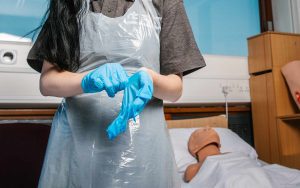 In a hospital room environment, a training figure model can be seen in a bed covered in white sheets with its head tilted back onto the pillow. Closer to us, a student is putting on a pair of blue latex gloves and wears a protective white plastic overall