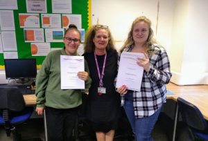 2 students holding certificates smile as they stand either side of a member of staff. All three smile towards camera, with a classroom setting behind them.