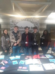 A group of 5 students stand in front of a Doncaster College banner, with a table containing books and promotional items in front of them