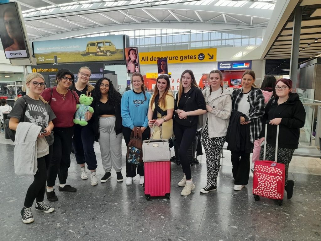 A group of ten students stand in a line smiling and posing in an airport terminal building. Two of the studens hold bright pink and red suitcases ready for the trip abroad.