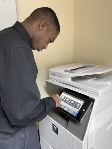 Student Marcel stands in front of a large waist-height photocopier machine, pointing at an opting on a touch screen display panel on it