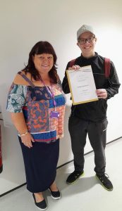 A student stands smiling proudly with a member of staff, holding up a certificate for participation in a work experience programme