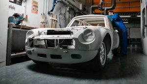 A large car chassis coated in white undercoat paint sits full framed with a student wearing blue overalls working on the back side of the car. The car is in a well lit garage environment