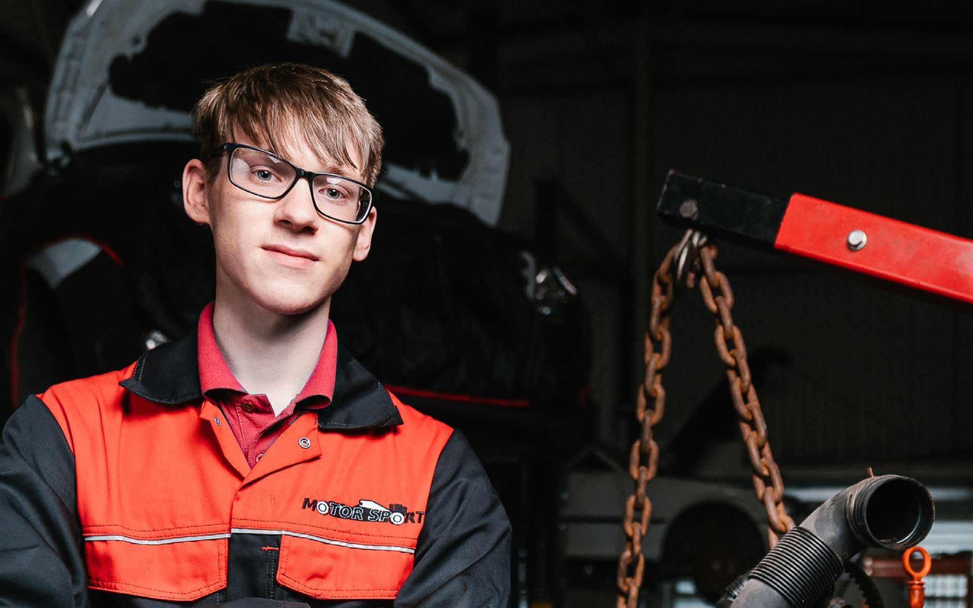 Stood in front of an open car bonnet, which is raised on a lifting platform in the garage workspace, student Jake looks intently at us with arms folded, wearing vibrant red and black workshop overalls. To Jake's side is part of some piping material and a harness chain holding some equipment that is out of view