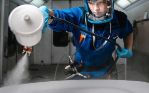 In a brightly lit enclosed workspace, a student wearing protective blue overalls and large clear face covering is holding a piece of paint spraying equipment in one hand, holding the tube that feeds it out of the way with the other hand. From the sprayer we can see a mist-like spray being delivered from the spray gun in a white or very light grey colouring