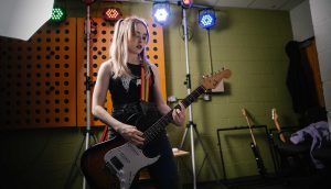 In a music studio room with soundproofing on the walls behind her and a large, upright lighting ring pointing towards us, a student stands proud playing a guitar