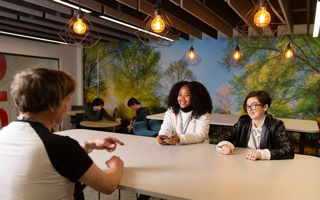 Sat around a large rectangular table, a student facing away from us is excitedly gesturing and telling a story to two other students sat opposite, facing us, who listen intently and smile. Behind them more students sit chatting, and on the rear wall is a large, bright picture of a bright blue sky and vibrant green trees. Industrial style lightbulbs hang decoratively from the ceiling around the room