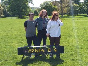 A group of three students stand together smiling in a large open landscape, with trees around 100 metres in the distance behind them. On the floor at their feet is a large digital scoreboard showing the results of one of their activities