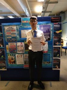 Student Tobias stands proudly holding up a certificate towards us, in front of a large display board containing promotional posters