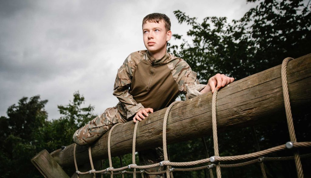 A student looking past us into the distant straddles on top of a climbing wall. They are laid over a large wooden beam with ropes visible draped over it for climbing.