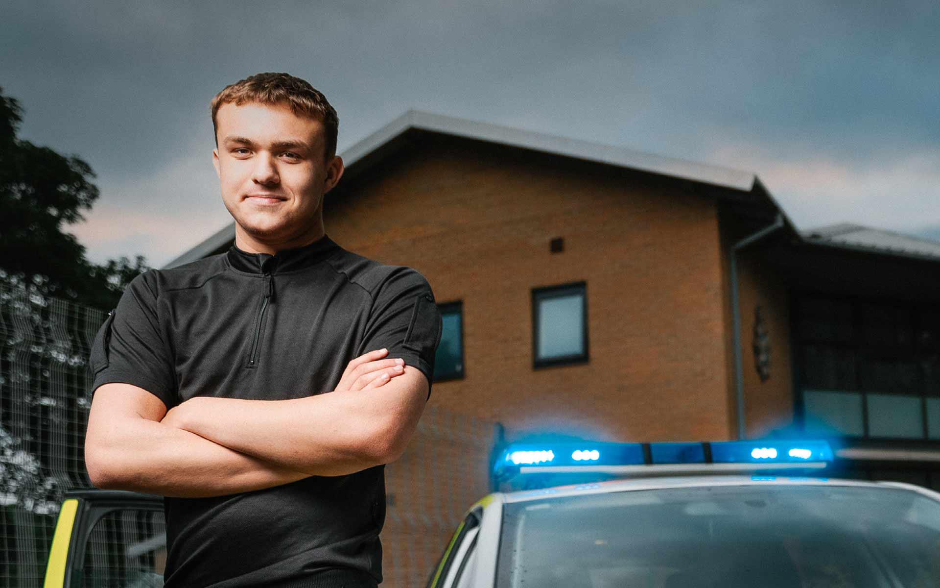Stood with arms folded and smiling toward us, Kacper poses in front of a police station building. Close behind him, the top of a police car with blue lights glowing is parked with the driver side door open
