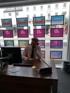 Sat at a wooden desk behind a computer, a smartly dressed student is talking on a telephone. He is sat in a property agency, with a large bright window behind displaying lots of promotional messages and information