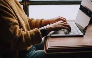 Seen from a side profile view, wearing a yellow jumper, a person sits at a wooden table typing at a laptop computer. A bright window behind them casts light towards us.