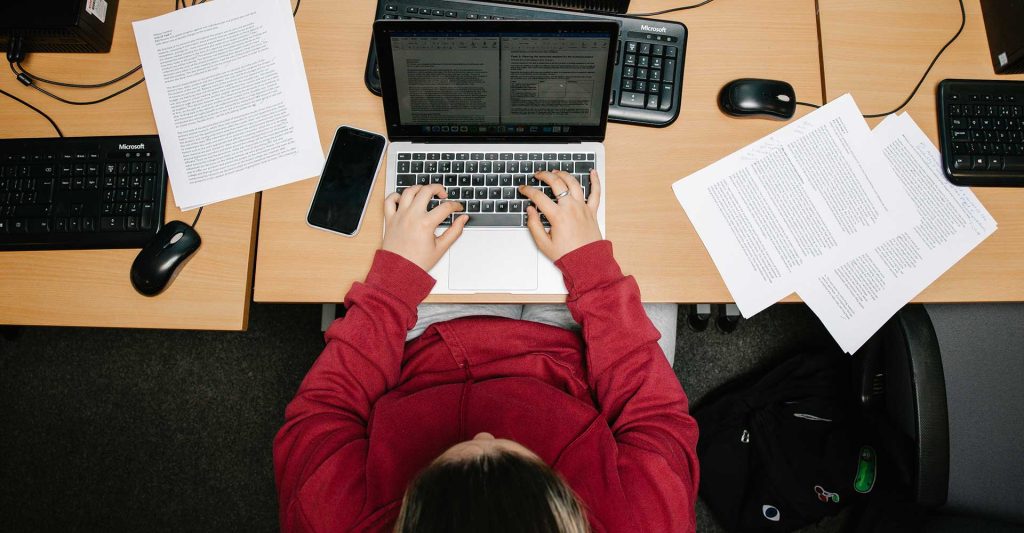 A view from above, we look directly down on a student wearing a red jacket typing at a laptop computer on a table. Surrounding the laptop are various notes on sheets of paper.