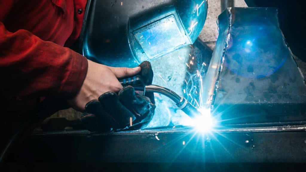 A person looks closely through a heavy duty welding mask while performing welding work on a tall tapered metal object in front of them. Sparks are emitted from the glowing welding torch
