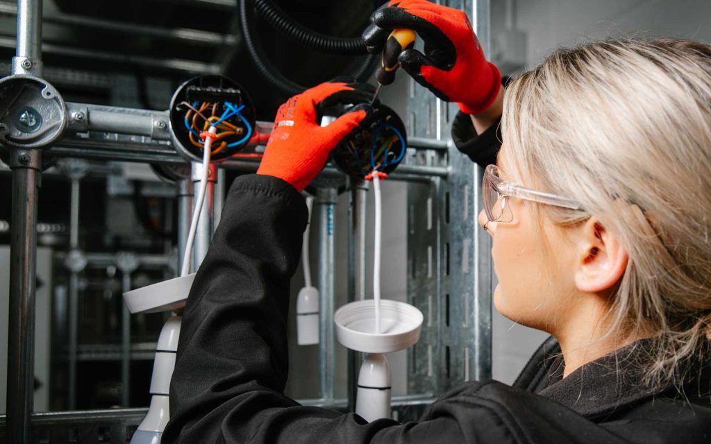 A student wearing protective clothing and gloves concentrates while working on some wiring held in front of them