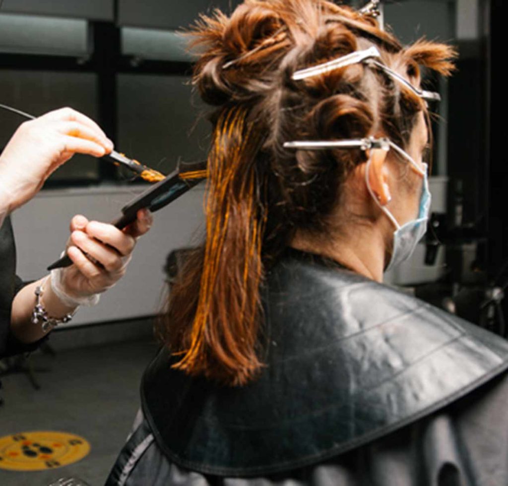 A student wearing gloves and mask holds a strand of another persons shoulder-length hair with one hand, the other using a brush to apply hair colouring to the hair. The rest of the person's hair is pinned and clipped up and around her head as the hairdresser performs the work