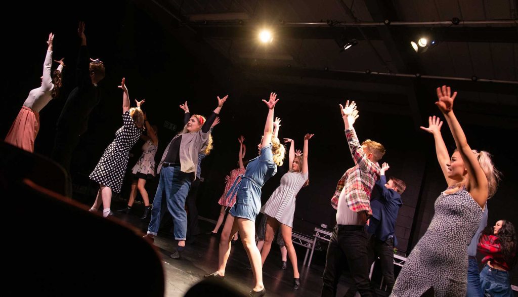 On a dark stage with a single spotlight above them, a large group of performing arts students reach up as if reaching for the light.