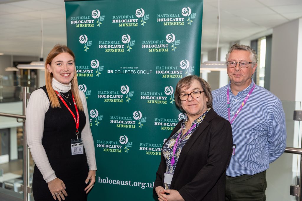 Three people stand in a bright indoor atrium in front of a green National Holocaust Museum display banner featuring repeated museum logos and the text “In partnership with DN Colleges Group.” All three wear staff lanyards and face the camera. Glass railings, hanging lights, and upper-floor walkways are visible in the background.